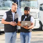 Two truck drivers reviewing fleet management data on a laptop, standing together in front of semi trucks