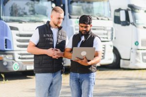Two truck drivers reviewing fleet management data on a laptop, standing together in front of semi trucks